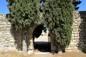 Zugang durch den Torbogen in der Sandsteinmauer der links und rechts davon von zwei Bäumen flankiert wird am Filerimos Kloster auf der Insel Rhodos. Beim Blick durch den Torbogen erkennt man die Ruinen von noch einem älteren byzantinischen Gebäude im Hintergrund.