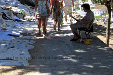 Eine Rhodierin die am Weg zur Ritterburg in Lindos im Schatten der Pinien versucht gestickte Tischdecken an die Touristen beim Griechenland Urlaub zu verkaufen.