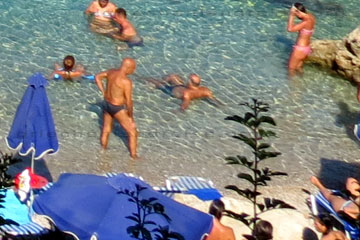 Braun gebrannte Urlauber am Strand auf der Insel Rhodos die sich im glasklaren Meerwasser vergnügen. Im Vordergrund Badegäste in dieser schönen kleinen Bucht die ein Sonnenbad auf einer der Liegen nehmen die man sich zusammen mit dem Sonnenschirm mietet.