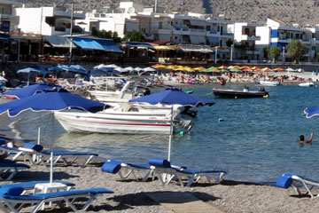 Kleinere Hotelanlagen direkt am Strand an einer kleinen Bucht auf der griechischen Insel Rhodos. Hier auf dem Bild zu sehen, der Kiesstrand, viele Sonnenliegen, blaue Sonnenschirme und im Hintergrund hinter dem Dorf eine senkrecht in die Höhe steigende Felswand.