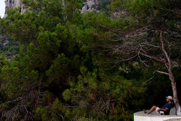 Ein Tourist der sich unter einem Schatten spendenden Baum auf einer Bank sitzt und die schöne Aussicht genießt. Im Hintergrund das satte Grün der Vegetation der Insel Samos, in Potami.