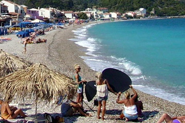 Strandleben auf der Insel Samos mit türkis farbenes Meerwasser in der Bucht. Sonnenschirme mit rundem Strohdach und Liegen am Kiesstrand und dem weißem schäumen der leichten Brandung.